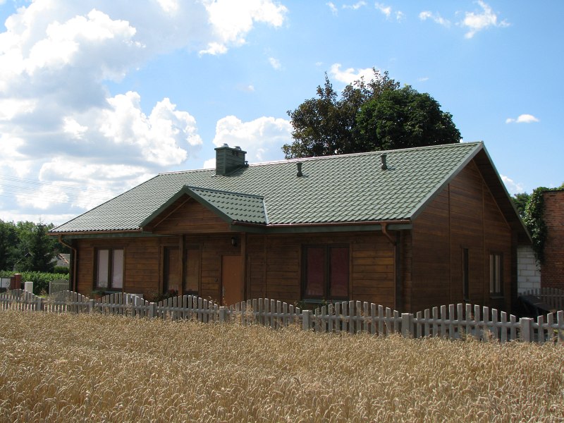 Parterowy dom z ciemnego drewna z zielonym dachem, otoczony niskim, jasnym płotem i polem zboża, widok z przodu w słoneczny dzień.